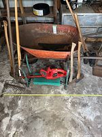 Photo showing rusty metal wheelbarrow, red electrical hedge trimmer, wooden-handled rake and other hand tools leaning against wall in a workshop setting.