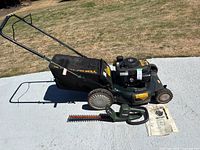 Overview of green Yard-Man push mower with attached cloth catcher and adjacent Black & Decker hedge trimmer on concrete surface