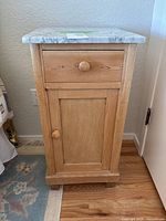 Front view of pine wood side table/nightstand with a single drawer and cabinet door, round wood knobs, set on wooden legs, placed on wood floor near a wall.
