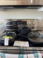View of entire set on stovetop showing 3 saucepans stacked, 2 frying pans with glass lids, large pot, and cooking instruction manuals.
