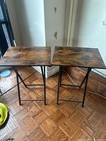 Two folding tray tables side by side on a wooden floor near a window, showing the full view including legs and tops.