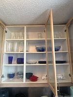 View of upper and middle shelves in cabinet showing clear glass carafes on top, blue glass bowls on upper right, multiple glass glasses and cups on left, ceramic bowls with lids and red ceramic pot on bottom shelf