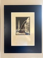 Framed monochrome photograph showing an urban fountain with obelisk and water spray at night, black wooden frame