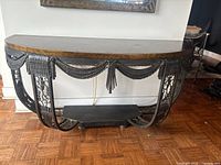 Front view of ornate console table showing black metal curved legs with garland and tassel decorations and wooden top with a gold mottled finish.