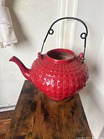 Front and side view of a red ceramic teapot with a textured geometric pattern and a black metal handle, sitting on a wooden surface.