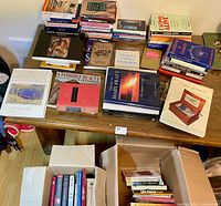 Tabletop view showing multiple stacks of books plus two open boxes of additional books