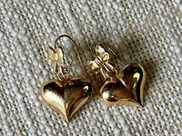 Pair of yellow gold earrings with bow and heart dangle photographed on textured beige fabric.
