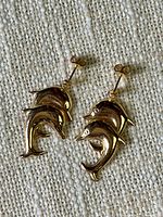 Pair of polished yellow gold dolphin dangle earrings with post backings. Showing front side view on fabric background.