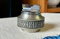 Pewter table lighter with decorative band and chrome ignition top shown on white cloth background on a wooden table.