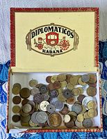 Open cigar box labeled Diplomaticos Habana containing a mixed collection of coins in various metals and sizes spread inside.