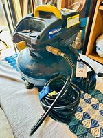 Full view of Mastercraft air compressor showing blue tank, yellow handle, black control panel, pressure gauges, and coiled air hose attached.