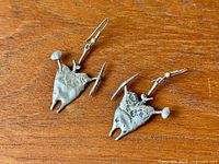 Pair of sculpted sterling silver earrings laid on wood surface, showing abstract anthropomorphic shapes with antler-like protrusions and hammer-like ends.