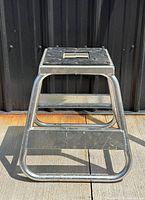 Front view of the modernist aluminum motorcycle stool showing its sturdy tubular design and square top with cutout handle.