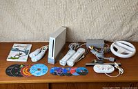 Full overview of white Wii console on stand, two nunchuks, two silicone-sleeved Wii Remotes, AC adapter, composite AV cable, sensor bar, steering wheel and row of discs