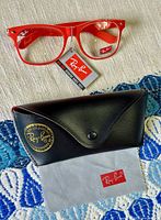 Red frame Ray-Ban Wayfarer eyeglasses with black case and cleaning cloth on patterned surface.