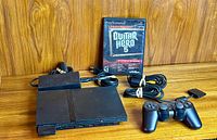 PlayStation 2 Slim console with controller, Guitar Hero 5 game disc case, cables, and memory card arranged on wood surface.