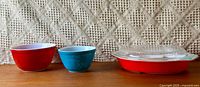 Three Pyrex bowls and a red lidded Pyrex casserole dish with glass lid displayed on wooden surface with textured beige background