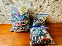 Three large resealable plastic bags filled with a mix of quartz watch cases, straps, and components in various colors and styles laid on a wooden surface against a knit fabric background.