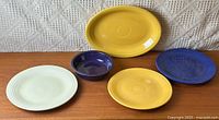 Five pieces of vintage Fiesta Ware arranged on a wooden surface: two yellow plates including an oval platter, a blue plate, an ivory plate, and a small purple bowl.