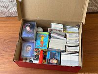 Photo showing a shoebox filled with multiple stacks of trading cards, including Star Trek, Simpsons, baseball and hockey cards plus a golf ball and coins visible inside the box.