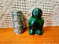 Photo of 4 cylindrical pewter napkin rings with carved West Coast Indigenous designs, stacked beside a carved green owlpik owl sculpture on textured fabric background.