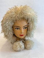 Front view of cream shaggy fur hat on mannequin head showing hood shape and pom-poms