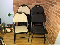Four folding chairs stacked against brick wall showing two beige and two dark grey fabric cushions on black metal frames