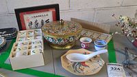 Overall lot on table showing framed Asian calligraphy print, floral porcelain napkin rings in boxes, colorful metal lidded bowl, small cups, spoon and plate with Asian motifs, wooden plate, and partial view of place setting name plates.