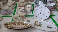 Photo of china items including floral teacups, saucers, and oval floral serving trays on a green-lined table.