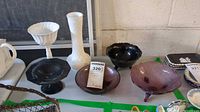 Wide photo showing several glass and ceramic bowls and vases on a table including black glass pedestal bowl, purple/red glass bowls, white milk glass vase