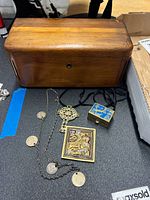 Top view of wooden Lane cedar box with costume jewelry and mini sewing kit arranged in front, showing wood grain and some scratches on box, several costume jewelry pieces including coin necklace, ornate cross pendant on black cord, framed saint pendant, and sewing kit.