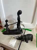 Photo showing two metal hiker figurines and one resin hippo figurine on display with background storage bin.