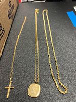 Three necklaces on black surface: cross pendant chain at left, monogram plaque chain center, rope chain at right