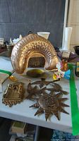 Overview photo of lot showing large brass fish wall hanging, sun-shaped metal decoration, religious plaque, basket and other items on table.