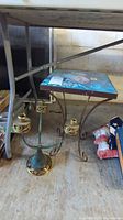 Photo of metal plant stand with painted top and brass five-arm candelabra on floor beneath a table.