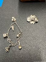 Photo showing sterling silver charm bracelet with five different charms arranged on a dark surface next to sterling silver flower pin with four heart-shaped petals.