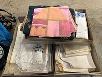 Box of assorted table linens including embroidered pink napkins, white lace-edged linens, and a blue textile piece.