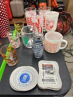 Front view of various vintage advertising glassware including Diet Coke tall glasses, souvenir shot glasses, and milk glass mug, along with two collectible ceramic plates showing advertising logos for Bookbinder's and a coffee brand.