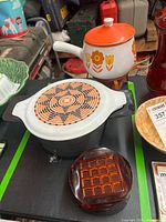 Overall view of vintage Pyrex ceramic casserole with lid, ceramic fondue pot with lid and stand, and amber plastic coasters.