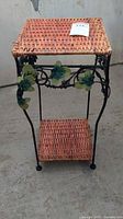 Front view of the wicker and metal plant stand showing the woven wicker top and bottom shelves with metal leaf accent details on the frame.