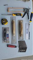 Overview showing tape measure, rasp, two finishing trowels, square trowel, taping knife, jab saw, mini hacksaw, caulk gun, coping saw