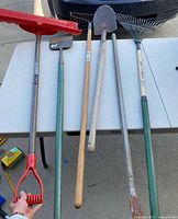 Five long-handled tools on table including snow pusher, tamper/hoe, shovel, rake
