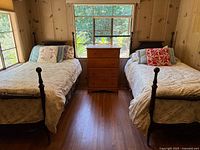 Pair of antique twin 4-poster beds in a wooden-paneled room with window behind, showing both beds side by side with bedding and pillows, wooden dresser in between beds but not included in lot.