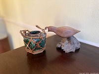 Photo displaying an antique dark green glazed small ceramic pot encased in woven rattan basket next to a carved wooden quail bird on a wooden base.