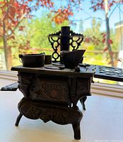 Side view of miniature cast iron vintage toy stove with detailed legs, chimney, and accompanying small pot and kettle.