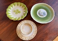 Lot of three antique decorative plates on wooden surface: two green scenic plates and one cream/gold floral plate.