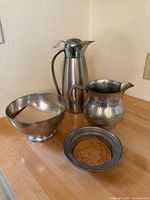 Four silver and pewter items on wooden table showing pitcher, two bowls, and coaster with cork