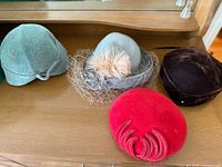 Four antique women's hats displayed on a wooden surface showcasing varied colors, materials, and decorative elements.