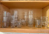 Wooden kitchen shelf with various clear glass cups and mugs, some stacked. Glasses vary in sizes and textures, some with handles and some without.