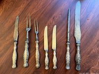 Full set of seven utensils laid out on wooden surface, showing ornate silver handles and varied blade and fork types.